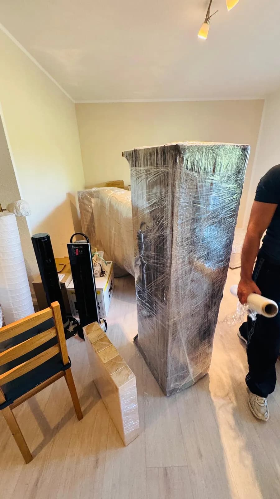 Hoher Schrank vollständig in Stretchfolie verpackt, daneben ein Mitarbeiter mit der Folienrolle in der Hand