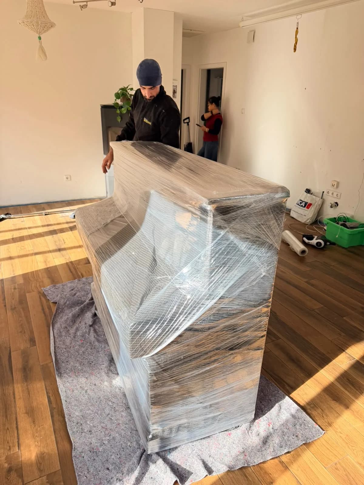 Klavier rundum gepolstert und in Stretchfolie gewickelt auf einem Vlies, das den Holzboden schützt, dahinter ein Mitarbeiter von Family Umzüge