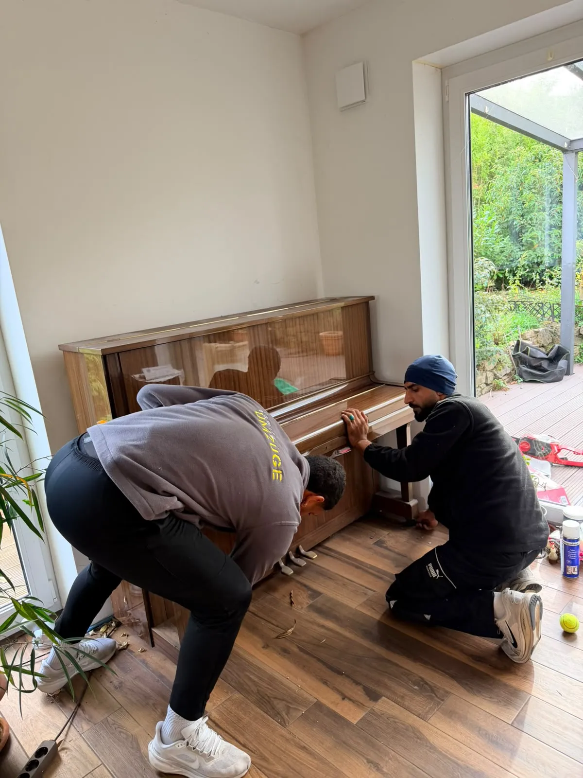 Zwei Mitarbeiter rücken ein Wandklavier von der Wand ab, einer kniet daneben und hält das Gehäuse an der Seite