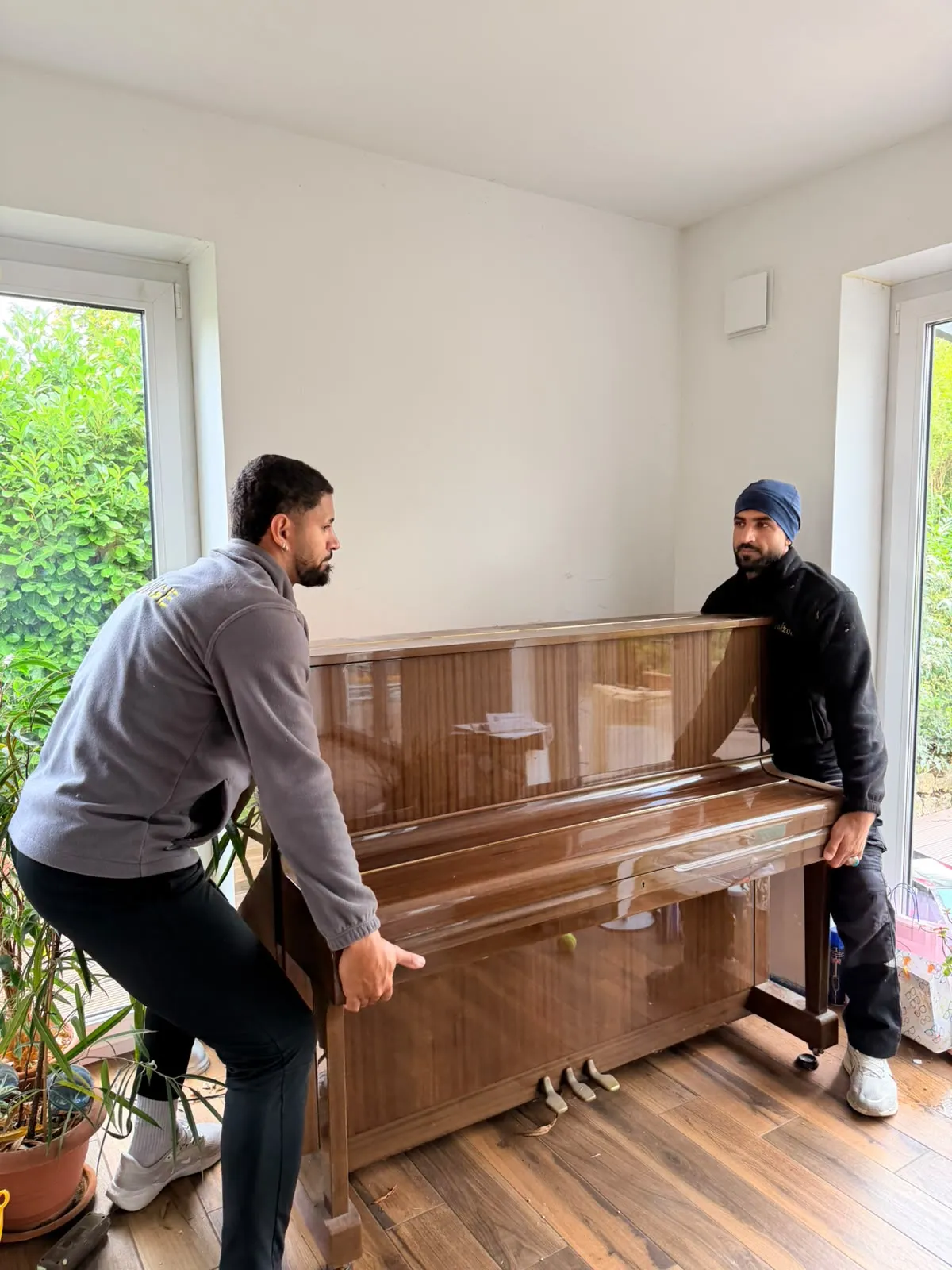 Zwei Mitarbeiter von Family Umzüge heben ein Wandklavier mit Nussbaum-Hochglanzgehäuse im Wohnzimmer an