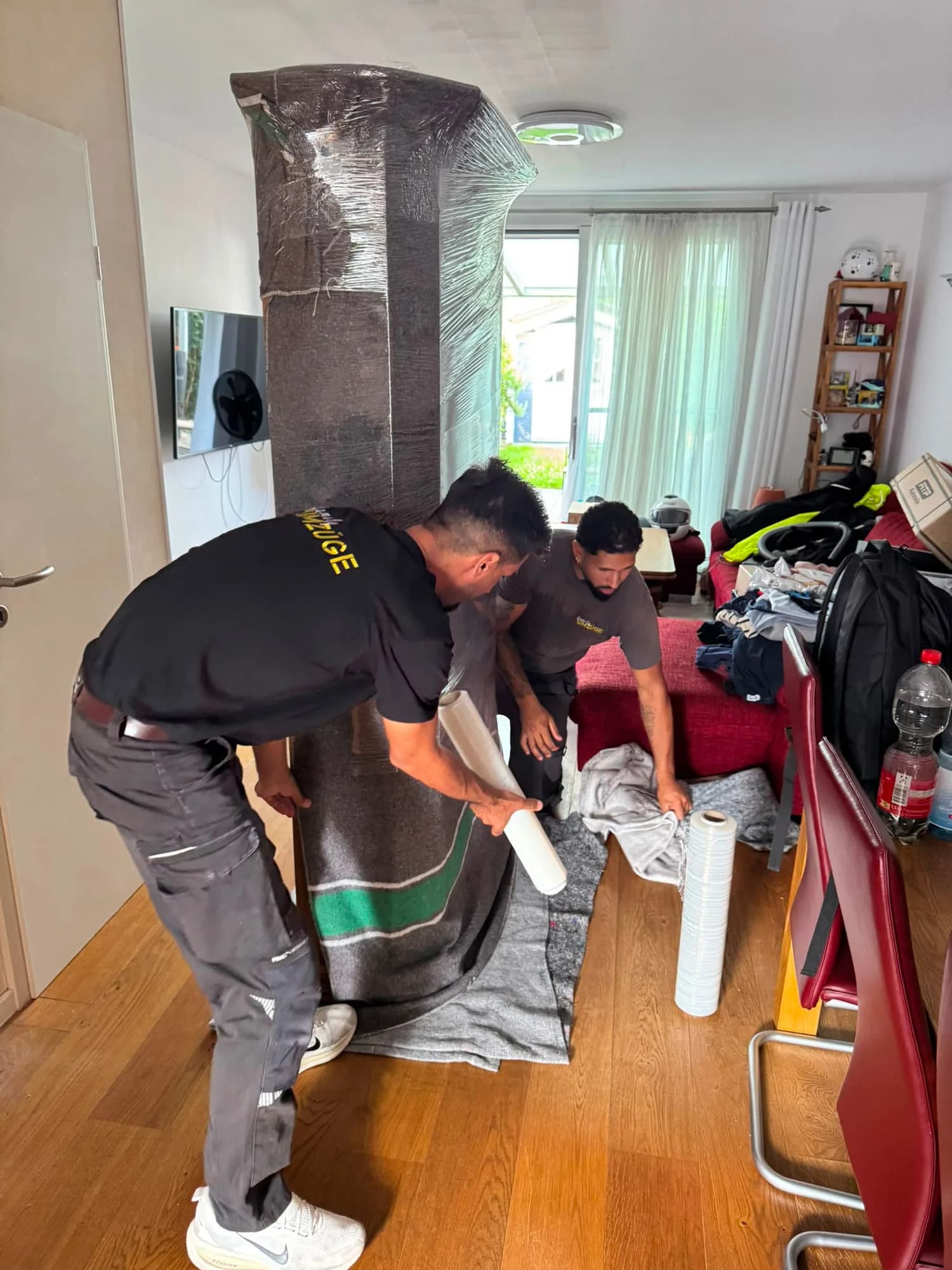 Zwei Mitarbeiter von Family Umzüge umwickeln einen hohen Schrank mit Stretchfolie und Umzugsdecke