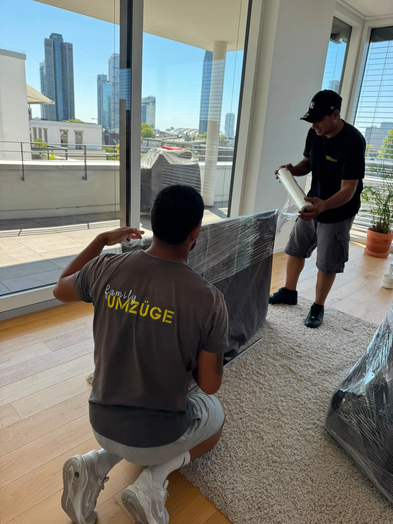 Zwei Mitarbeiter von Family Umzüge sichern ein Polstermöbel mit Stretchfolie – im Fenster die Frankfurter Skyline