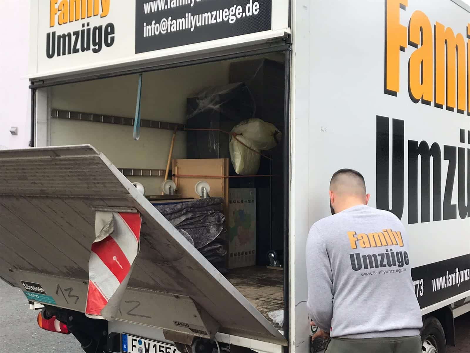 Ein Mann im Hemd von Family Umzüge lädt vorsichtig Möbel und Kisten in einen Markenumzugs-LKW mit geöffneter Heckklappe. Das Logo und die Kontaktdaten von Family Umzüge sind auf der Seite des Fahrzeugs zu sehen. - Family Umzüge